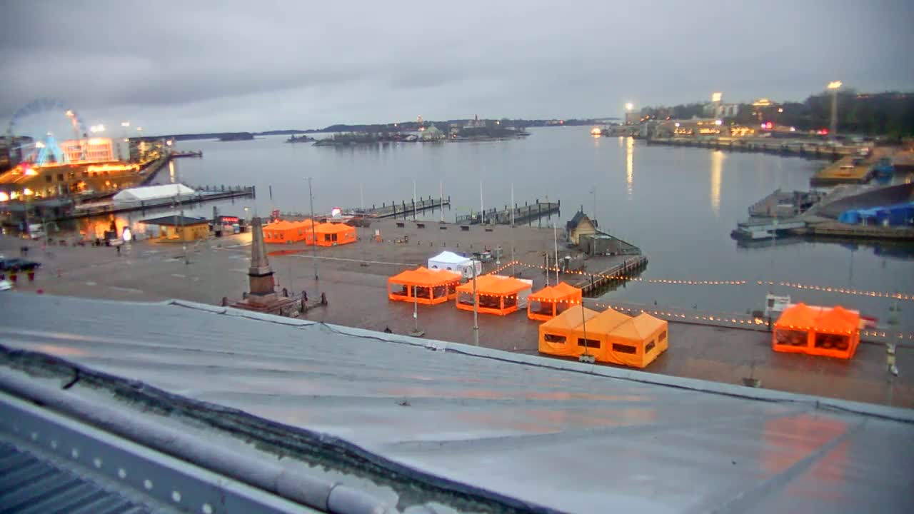 An overcast day reveals a harbor scene with several orange tents in a plaza near a body of water,  with buildings and docked boats visible in the distance.