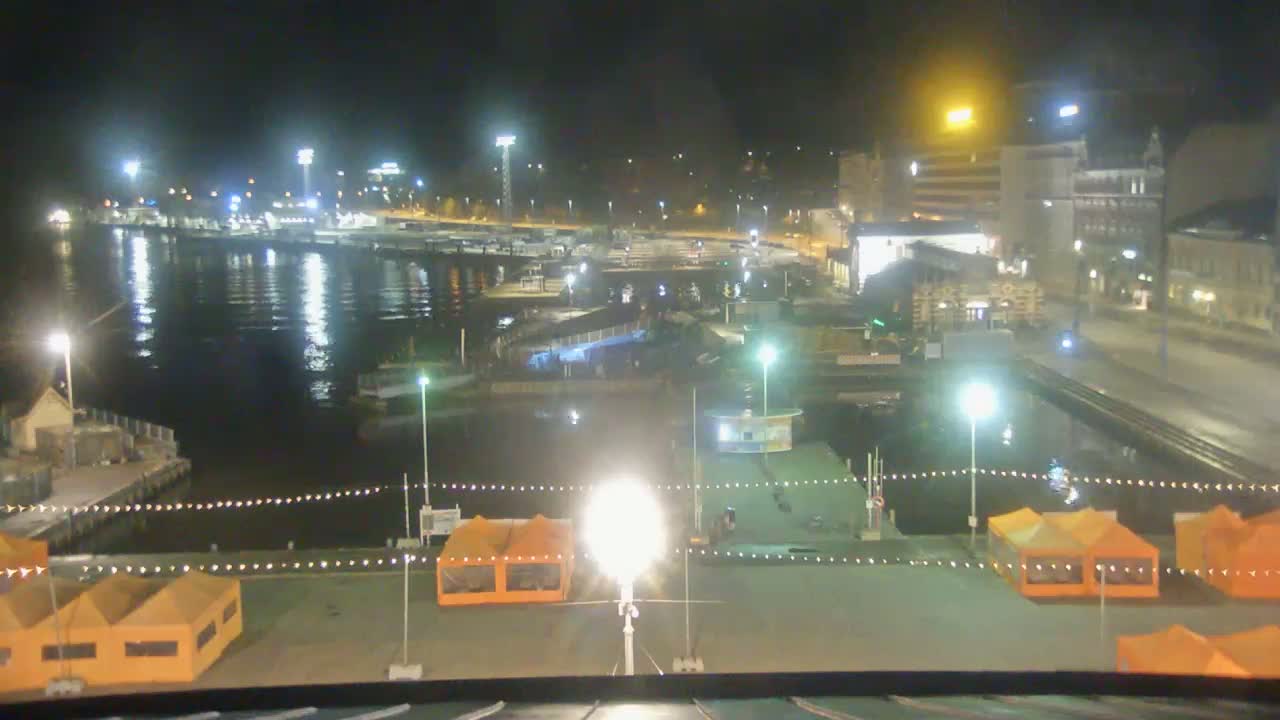 A nighttime view of a harbor with boats moored, buildings lit up along the shore, and several orange tents set up on a dock.