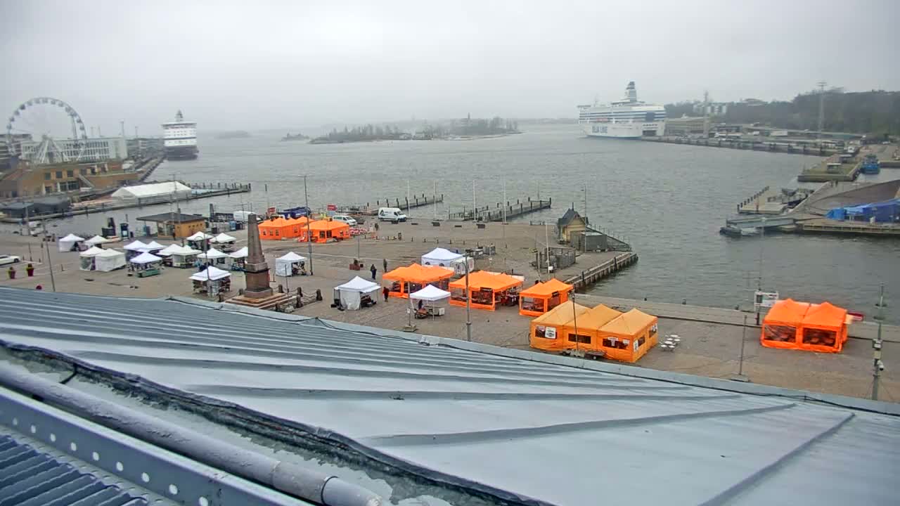 A harbor scene on an overcast day shows numerous market stalls set up on a waterfront plaza, with a Ferris wheel, ships, and buildings in the background.