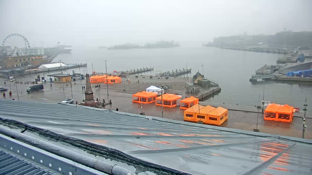 A foggy harbor scene shows numerous orange tents set up on a wet plaza near a waterfront with docked boats and a Ferris wheel in the distance.