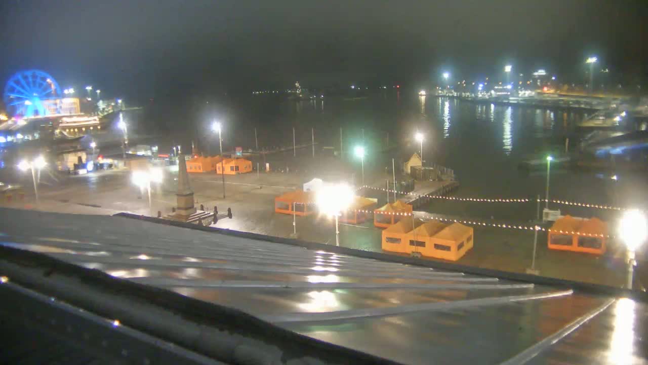 A nighttime view of a harbor with several orange tents, a Ferris wheel, and a monument, all illuminated under wet conditions.