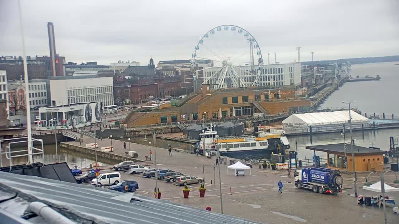 A Ferris wheel overlooks a harbor scene on an overcast day, with boats docked and cars parked in a lot near modern buildings.