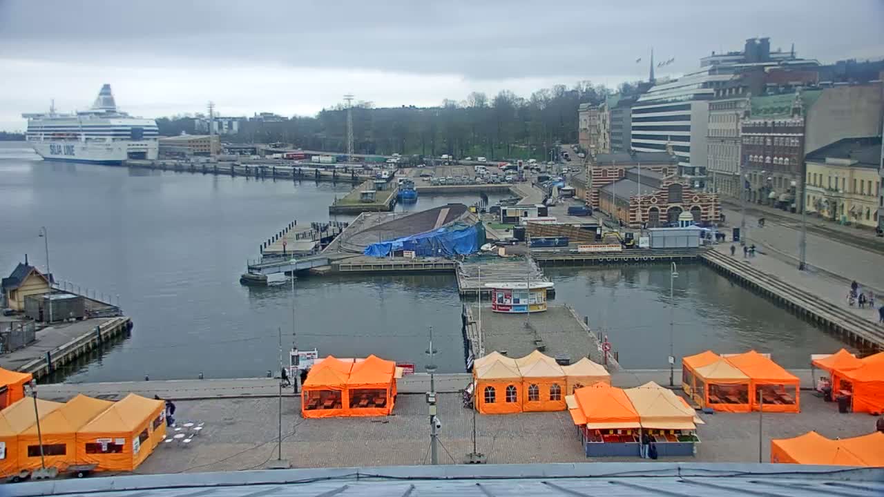 A harbor scene on an overcast day shows a large ferry docked, several buildings along the waterfront, orange market tents set up on a paved area, and a blue tarp covering something on a pier.