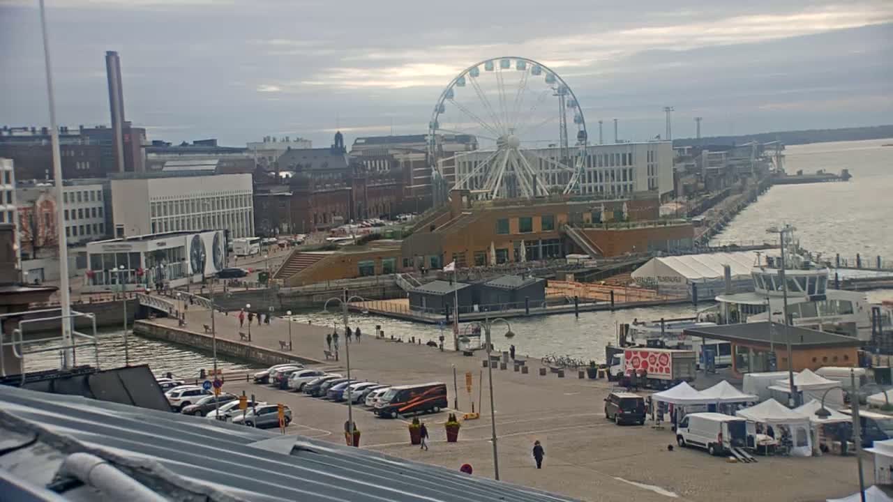 An overcast day shows a harbor city with a Ferris wheel, buildings, parked cars, a few people walking, and small market tents near the water.