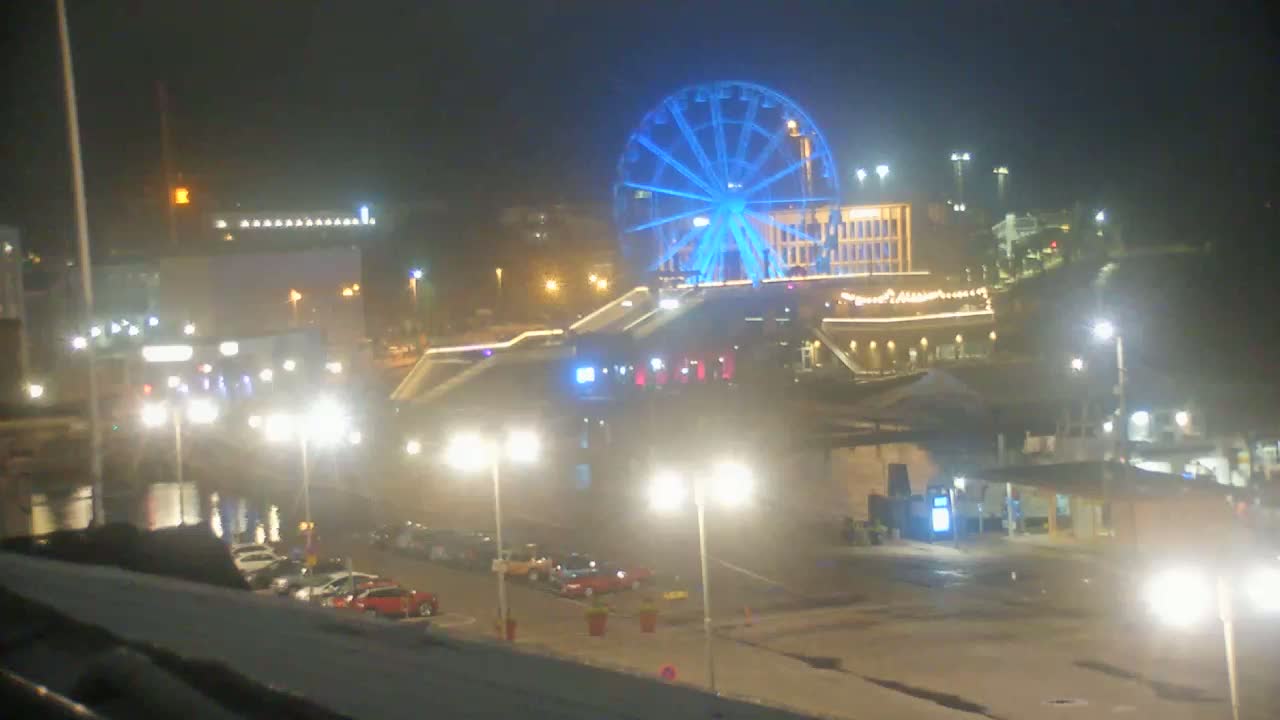 A brightly lit Ferris wheel glows blue at night next to a waterfront area with parked cars and streetlights in hazy conditions.