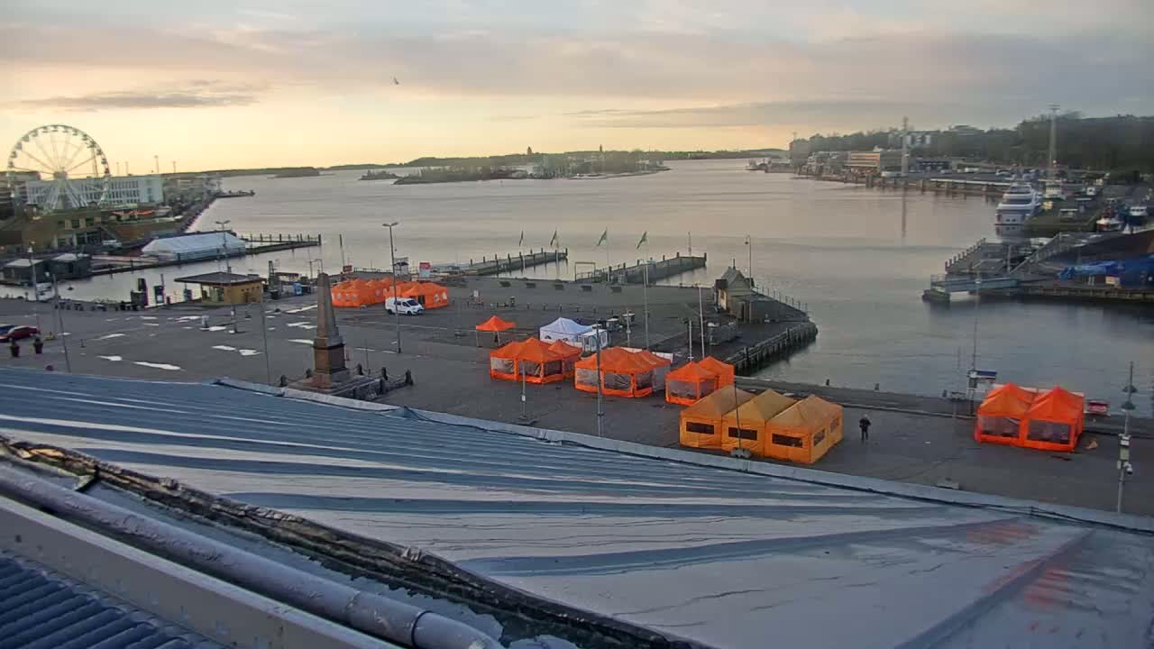 A harbor scene at sunrise shows numerous orange tents set up in a paved area next to a body of water with boats and a Ferris wheel visible in the distance.