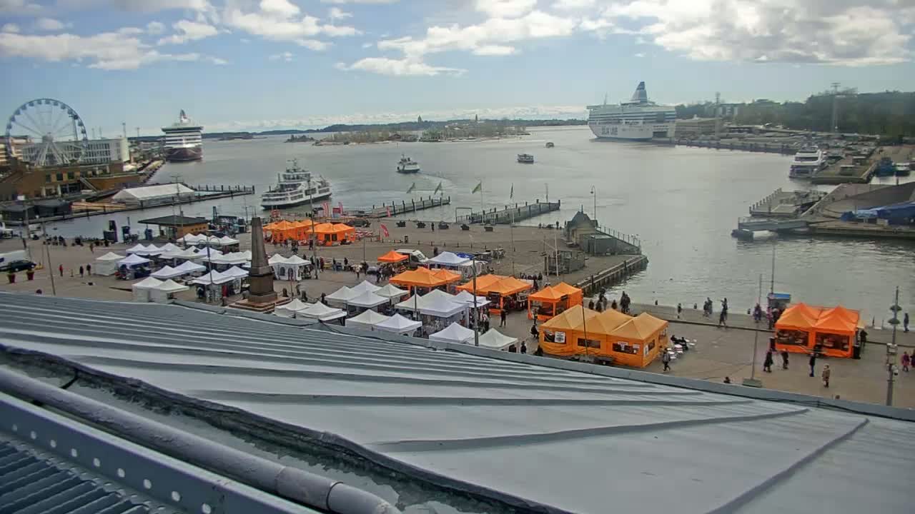 A harbor scene under partly cloudy skies shows a waterfront market with numerous white and orange tents, a Ferris wheel, several ships and ferries, and a calm body of water.