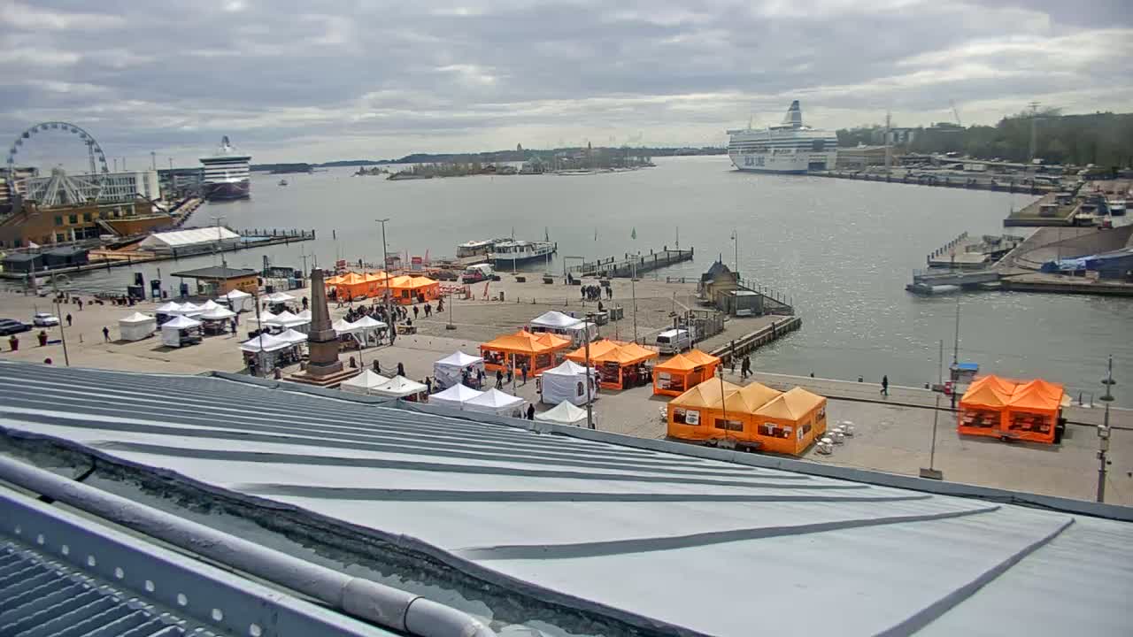 An outdoor market with numerous white and orange tents is situated on a waterfront plaza next to a harbor with several ships and a Ferris wheel under a cloudy sky.