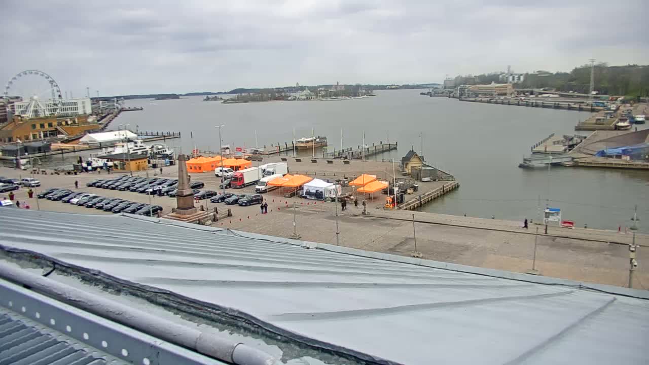 An overcast day shows a harbor with several boats, a parking lot full of cars, and orange tents set up on a paved area near a Ferris wheel in the distance.