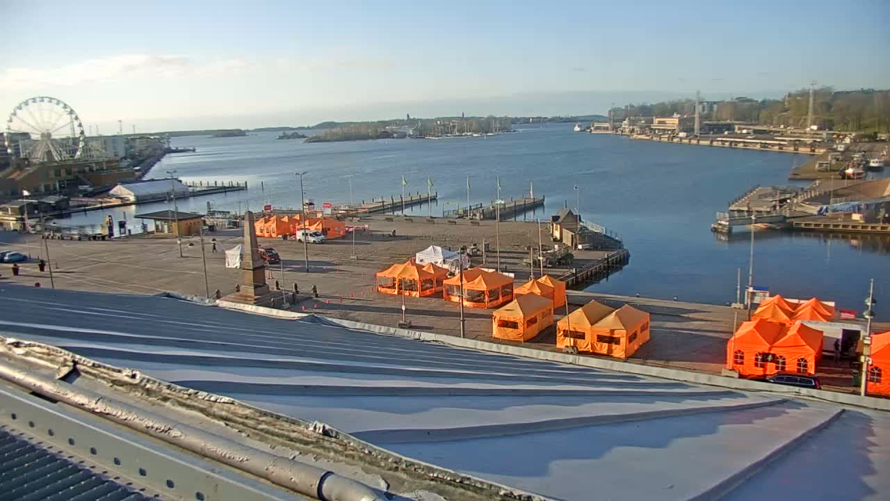 A harbor scene on a sunny day shows a waterfront area with numerous orange tents, a Ferris wheel, and boats docked, all viewed from a sloped metal rooftop.