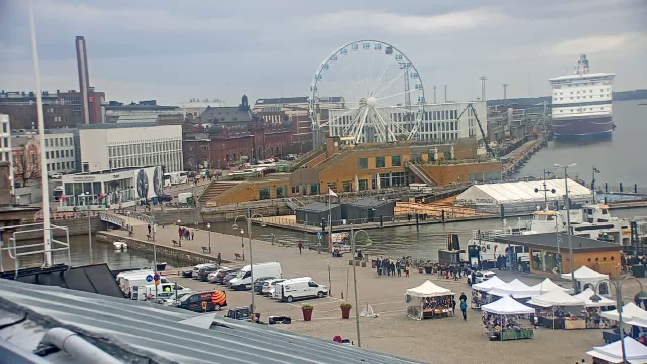 A harbor scene on an overcast day features a Ferris wheel, a large cruise ship, buildings, docked boats, cars in a parking lot, and market stalls.