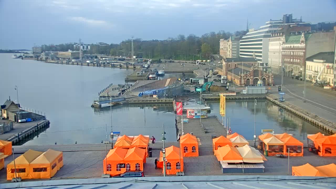 An overcast day at a harbor shows numerous orange tents set up on a dock next to calm water, with boats and buildings visible in the background.