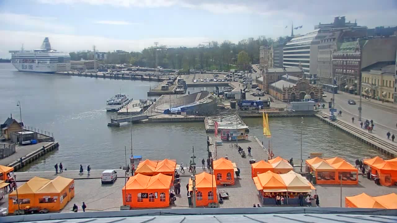 A harbor scene on a partly cloudy day shows a large ferry docked, smaller vessels in the water, a market with numerous orange tents set up on a dock, and buildings lining the waterfront.