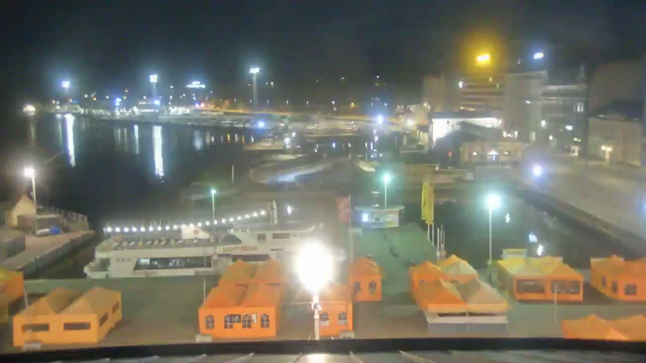 A nighttime view of a harbor with docked boats, orange structures, and buildings along the waterfront under clear skies.