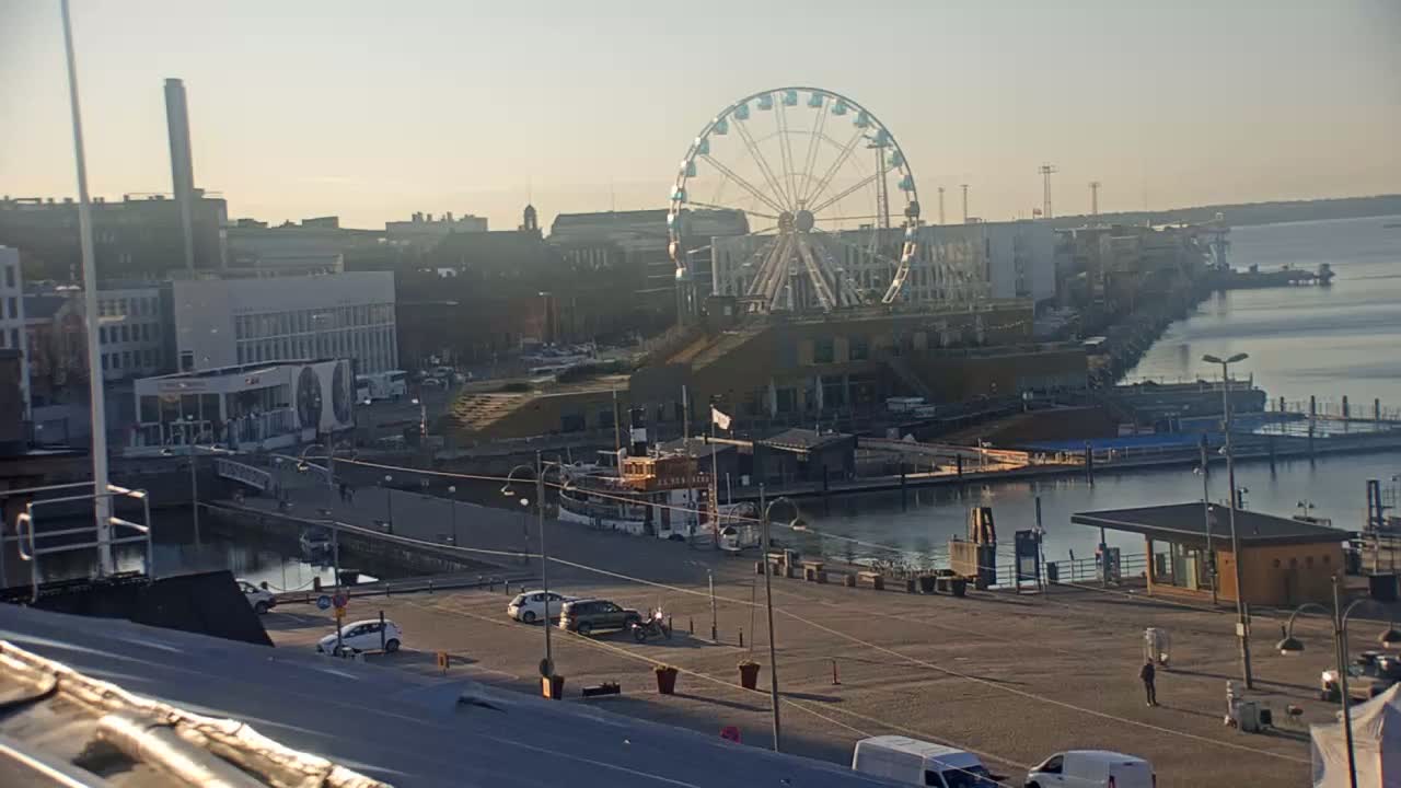 A Ferris wheel sits by a harbor on a sunny day, with cars parked in a lot and a small boat in the water.
