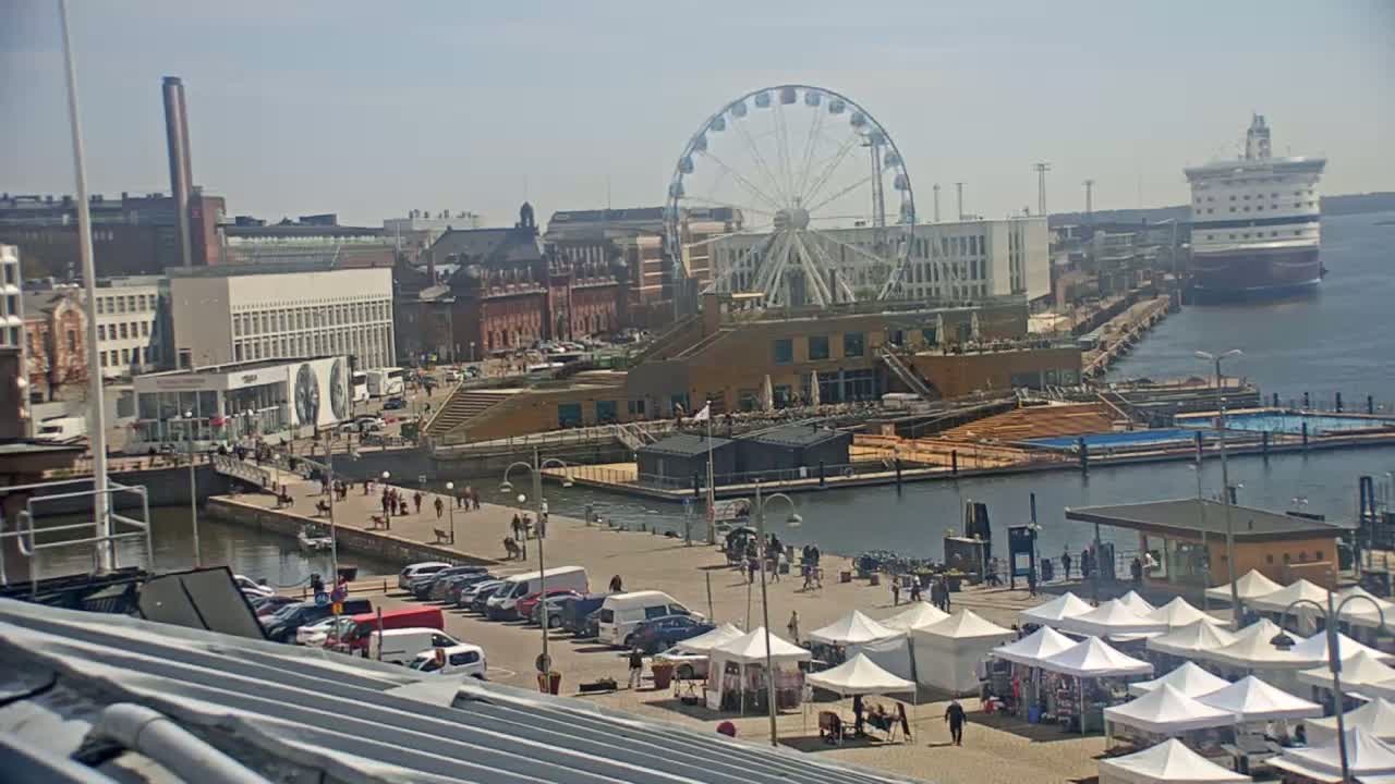 A harbor scene on a sunny day shows a Ferris wheel, a large ship, buildings, cars, market stalls, and people walking along a waterfront promenade.