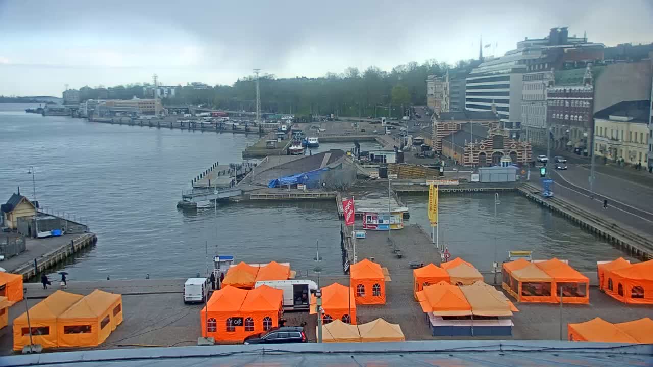 A harbor scene on an overcast day shows numerous orange tents set up on a dock, boats in the water, and buildings lining the waterfront.