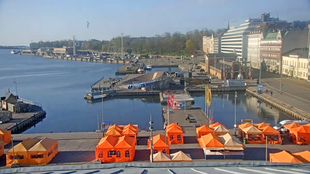 A harbor scene on a sunny day shows a calm body of water with boats, docks, buildings, and numerous orange tents set up on a paved area.