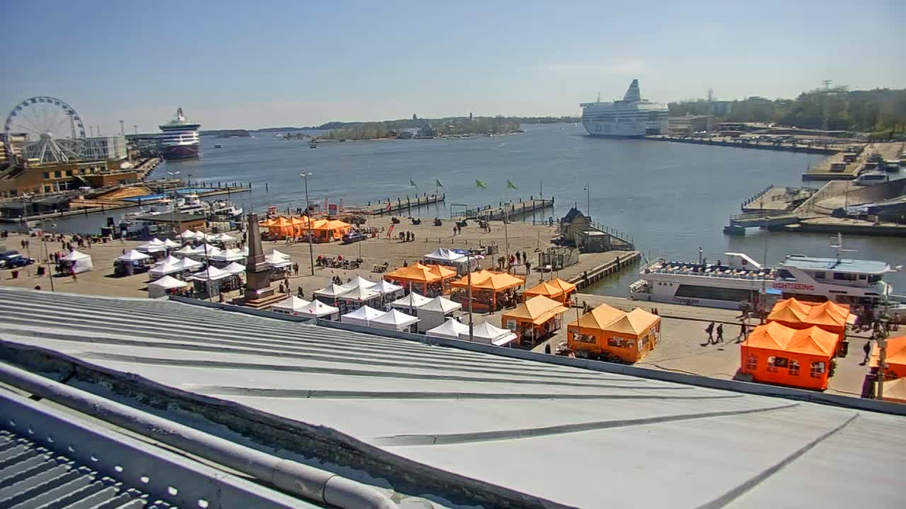 A harbor scene on a sunny day shows numerous market stalls, a Ferris wheel, several docked ships, and a passenger boat.