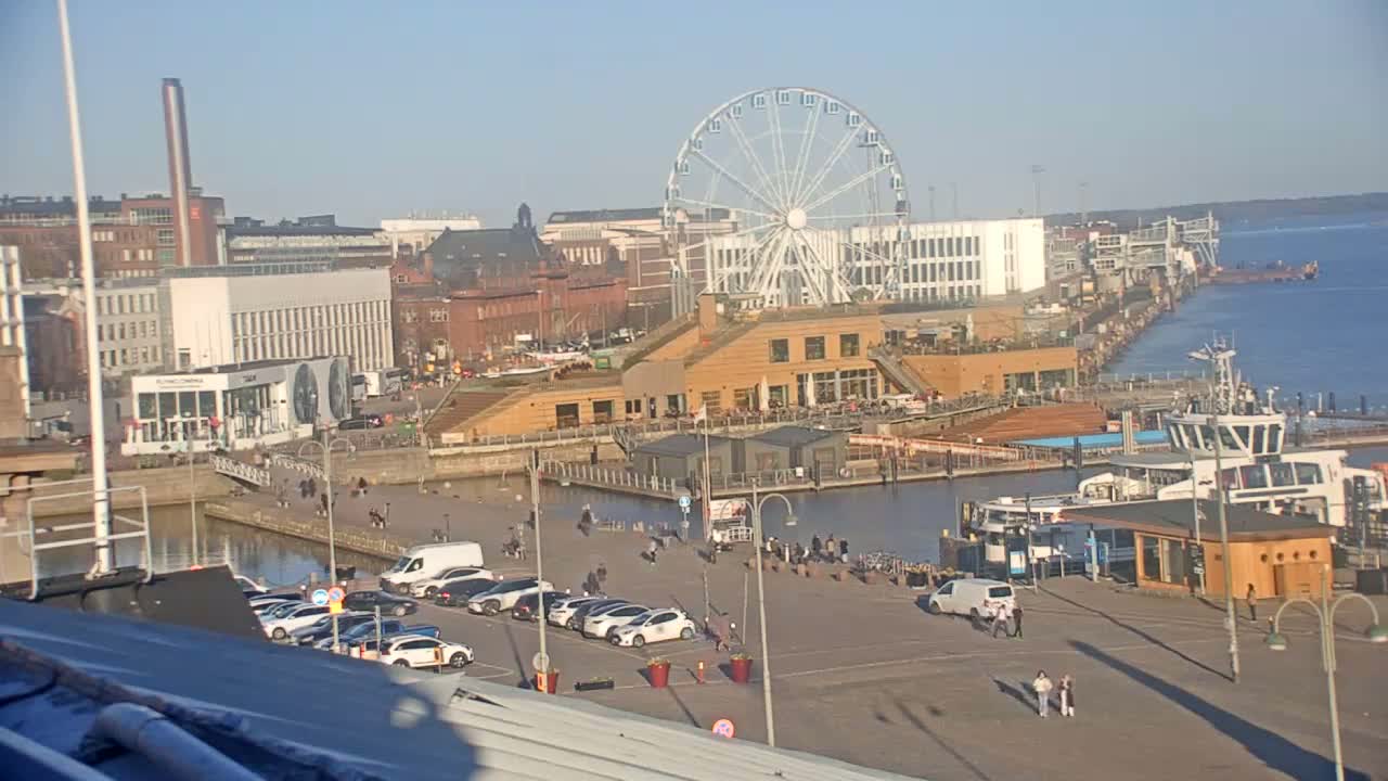 A waterfront city scene on a sunny day shows a Ferris wheel, buildings, docked boats, a parking lot with cars, and people walking around.