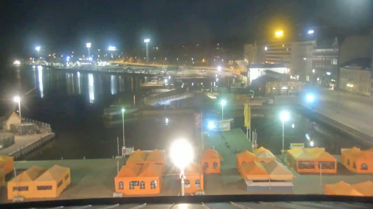 A nighttime view of a harbor with several orange tents, boats, and buildings illuminated by bright lights.
