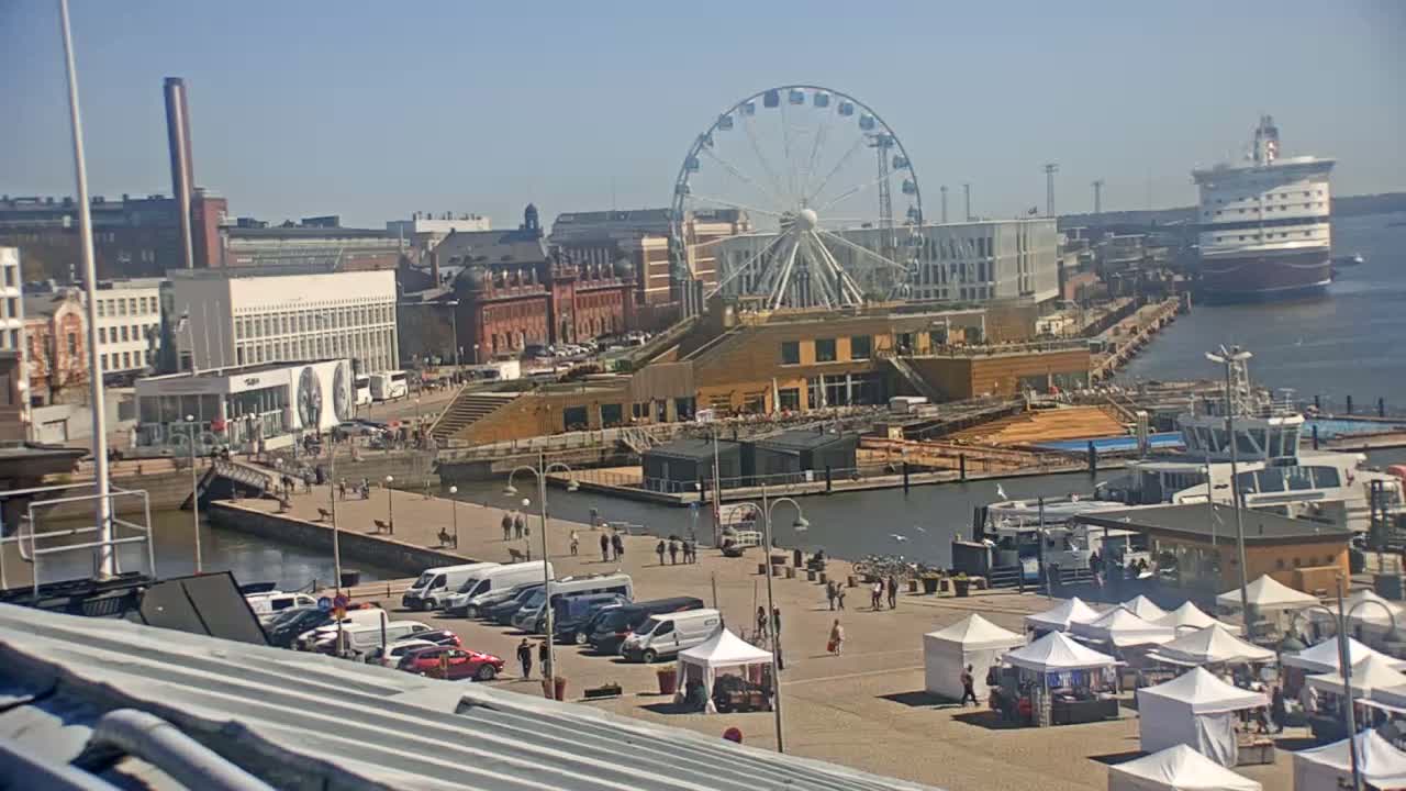 A harbor scene on a sunny day shows a Ferris wheel, buildings, boats, and a market with white tents.