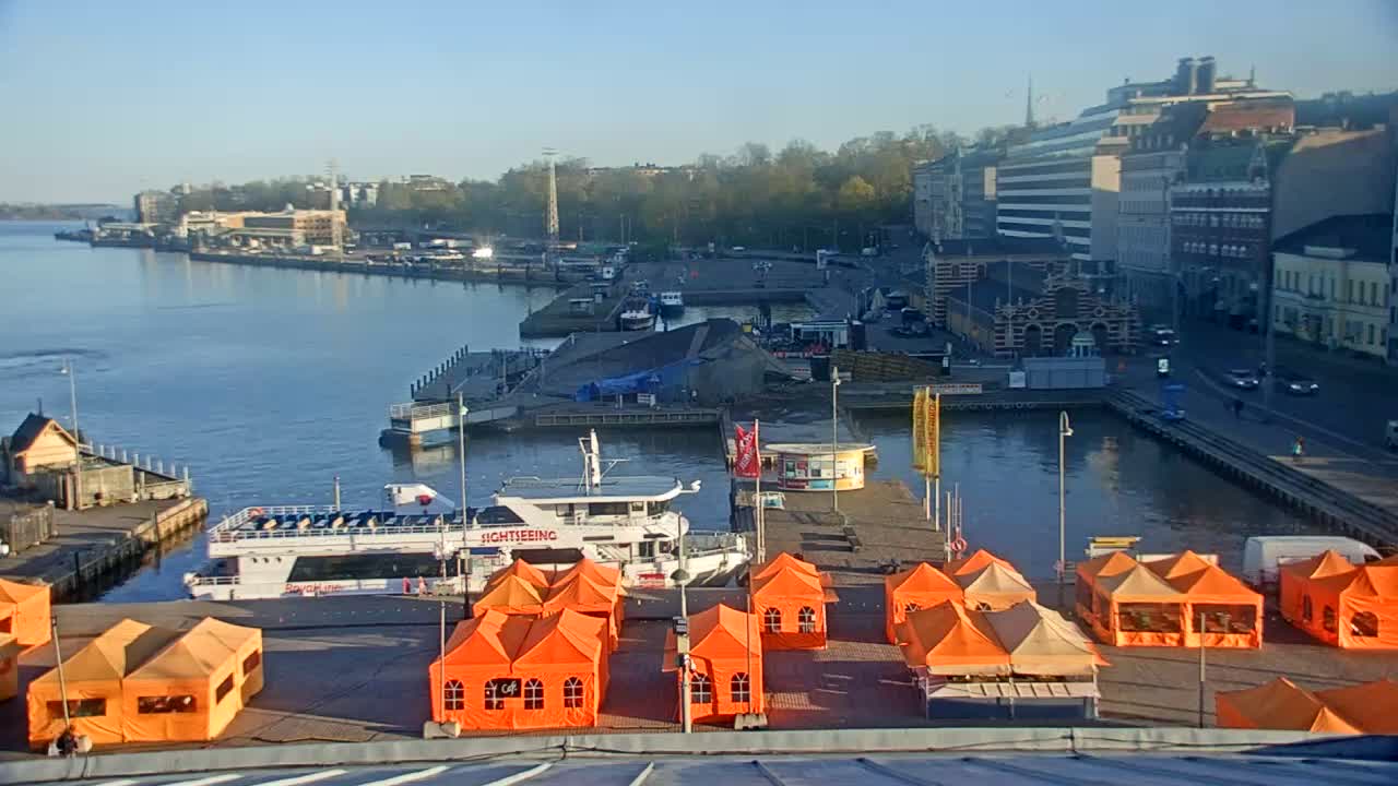 A harbor scene on a sunny day shows several boats docked near orange tents and buildings along the waterfront.