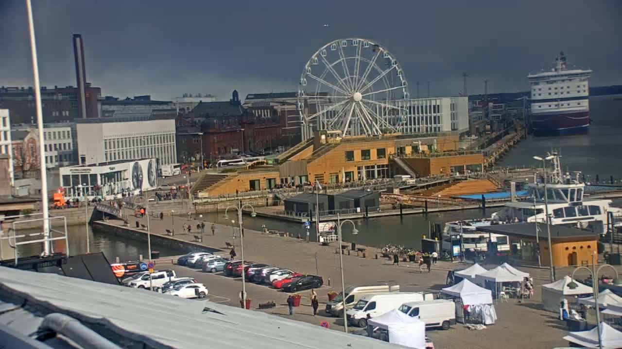 A harbor scene under partly cloudy skies features a Ferris wheel, a large cruise ship, several smaller boats, parked cars, and market stalls.