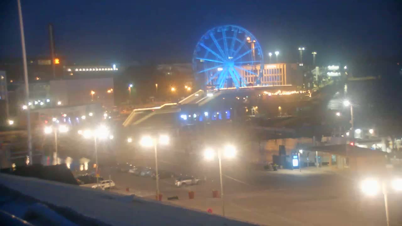A brightly lit Ferris wheel dominates a nighttime cityscape, with streetlights illuminating a parking lot and waterfront area.