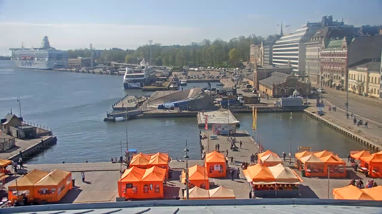 A harbor scene on a sunny day shows several docked ships, a city waterfront with buildings, and numerous orange market tents set up on a quay.