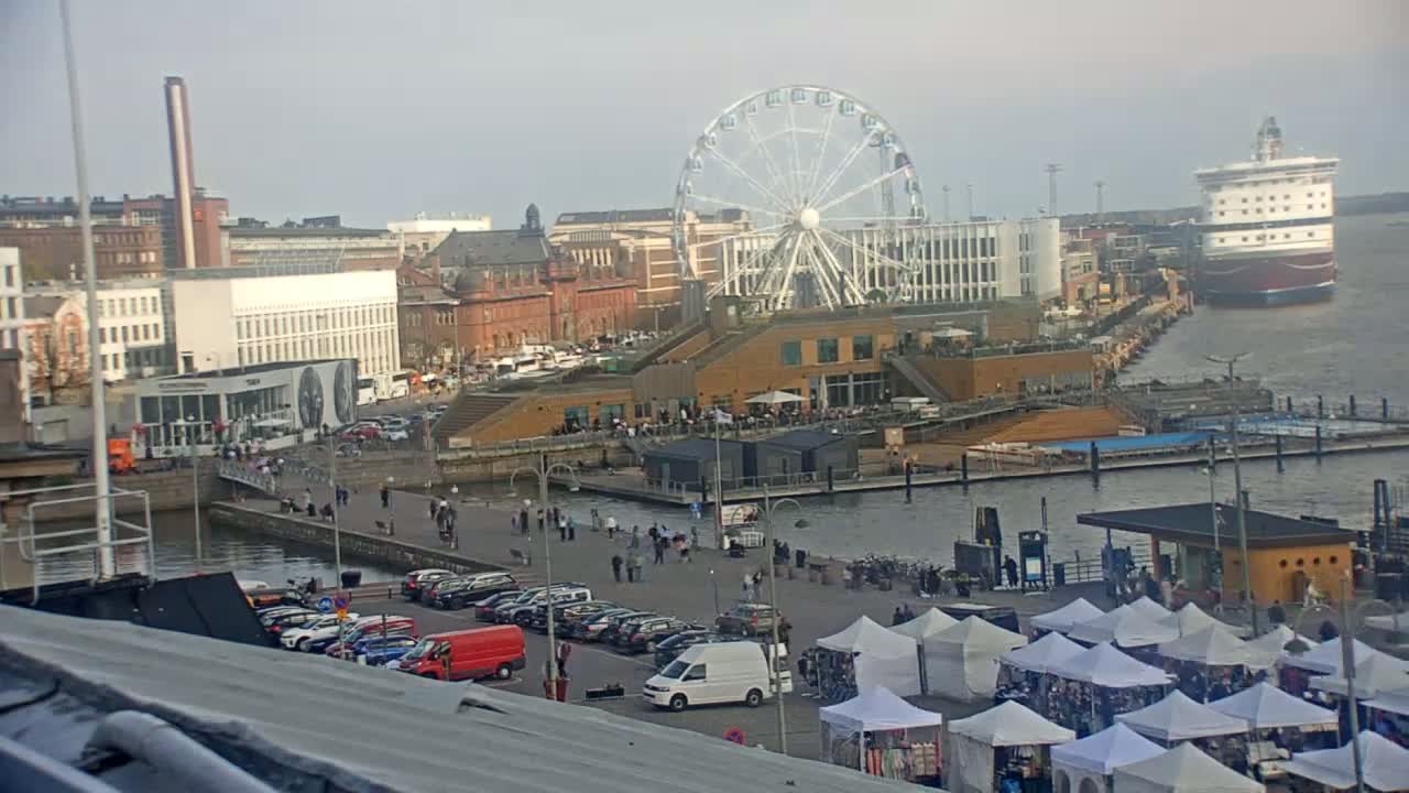 A waterfront city scene on an overcast day shows a Ferris wheel, buildings, docked ships, market stalls, and people walking along a harbor.