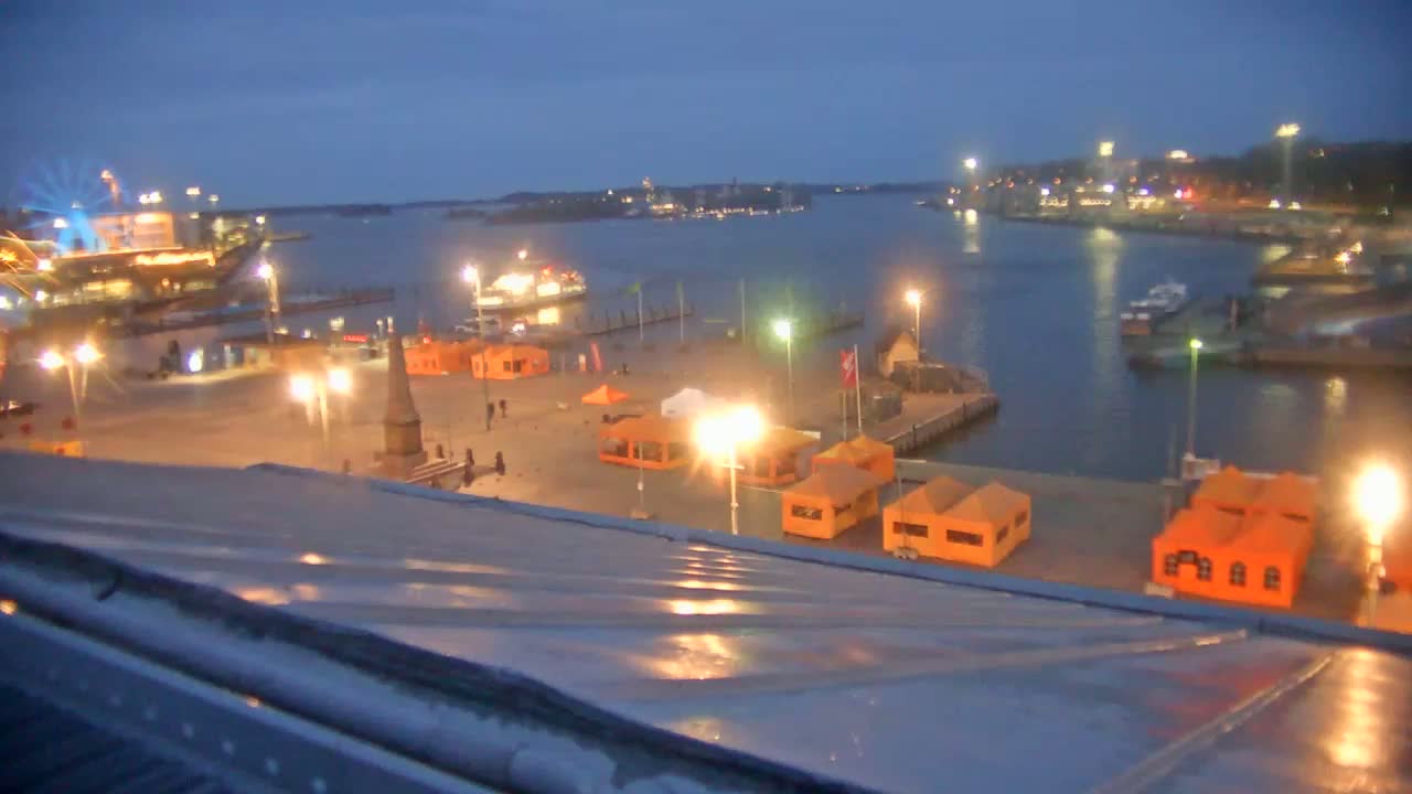 A nighttime view of a harbor with several boats and illuminated orange tents on a waterfront plaza, under a twilight sky.