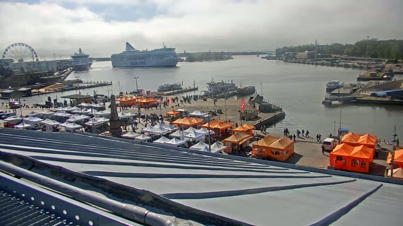 A harbor scene on a cloudy day shows numerous white and orange market tents set up on a pier near a Ferris wheel, with several ships in the water.
