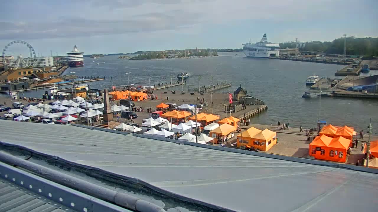 A harbor scene on a sunny day shows numerous market stalls, a Ferris wheel, and several ships in the water.