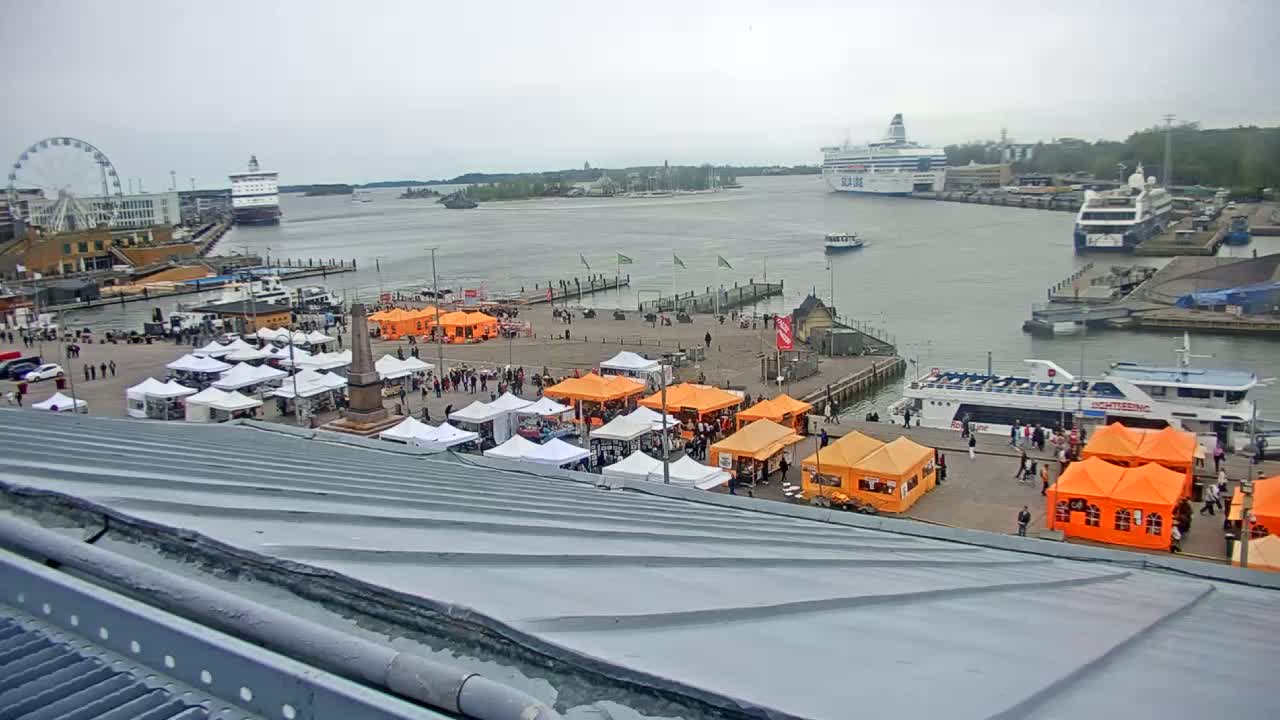 An outdoor market with orange and white tents occupies a harbor square, with ferries and a Ferris wheel visible in the background under an overcast sky.