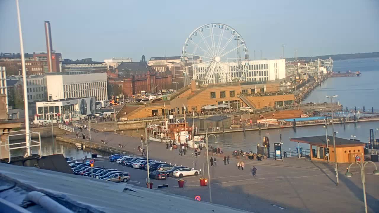A waterfront area with a Ferris wheel, buildings, a docked boat, and parked cars under a clear, sunny sky.
