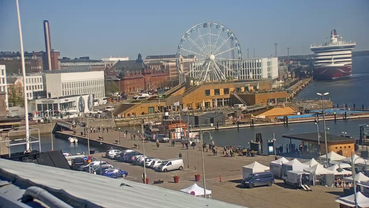 A waterfront city scene on a sunny day shows a Ferris wheel, buildings, a cruise ship in the distance, and numerous people and cars in a parking lot near market tents.