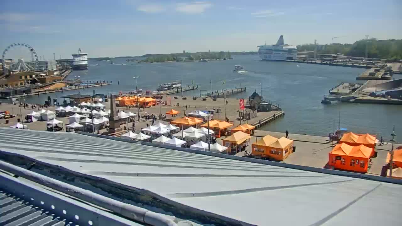 A harbor scene on a sunny day shows numerous market stalls and a Ferris wheel near a body of water with a ferry in the distance.