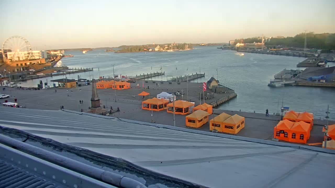 A harbor scene at sunset shows a large plaza with several orange tents, a Ferris wheel in the background, and calm water with boats and islands in the distance.