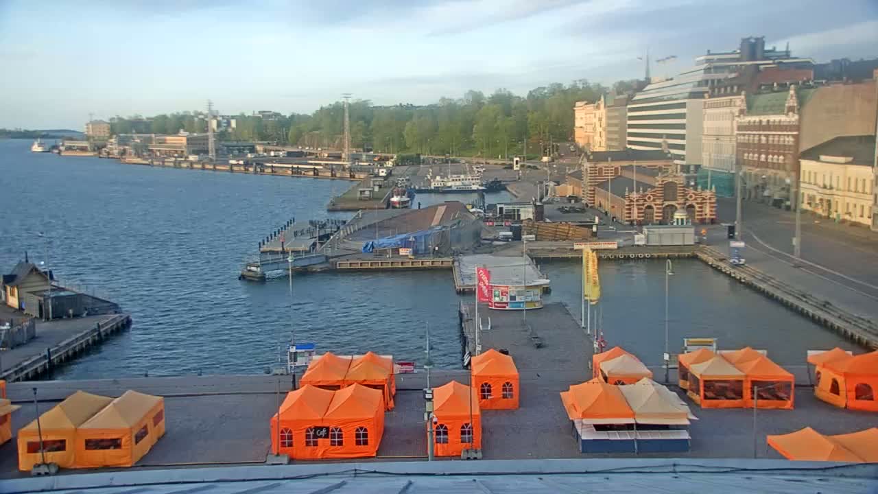 An overcast day at a harbor shows several boats docked near buildings, with orange tents set up on a pier in the foreground.