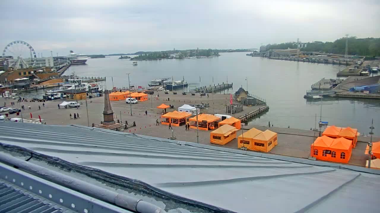 An overcast day shows a harbor with boats and a Ferris wheel in the distance, and a plaza with several orange tents.