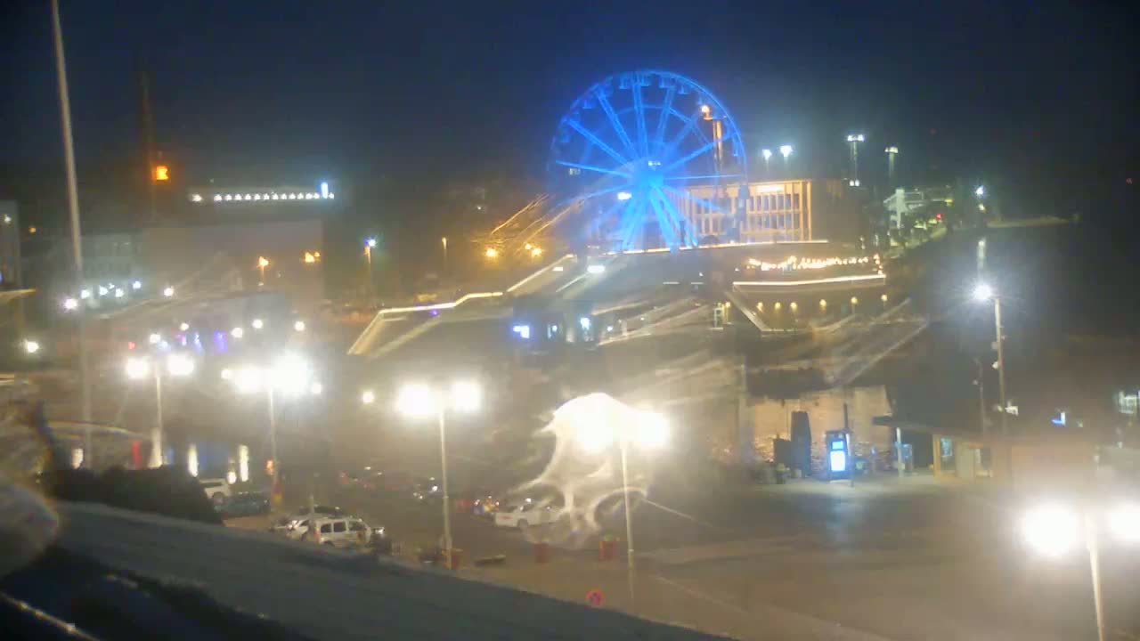A brightly lit Ferris wheel dominates a nighttime cityscape, with vehicles and streetlights visible in the foreground under hazy conditions.