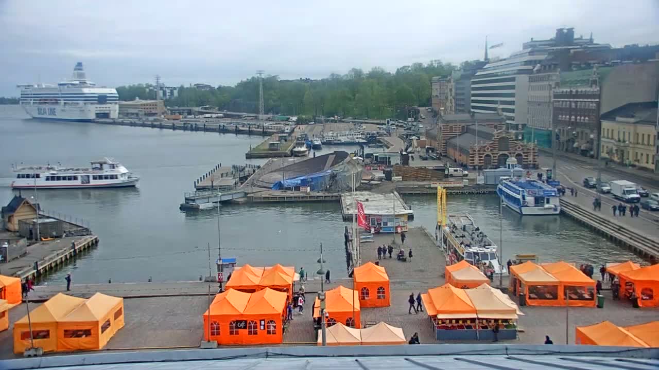 A harbor scene on an overcast day shows several boats, including a large ferry, smaller passenger vessels, and numerous orange market tents set up on a pier alongside buildings.