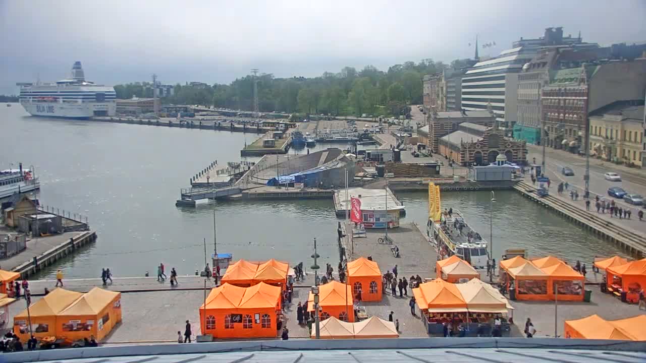 A harbor scene on an overcast day shows a large cruise ship in the distance, numerous smaller boats and docks, several orange market tents, and buildings lining the waterfront.