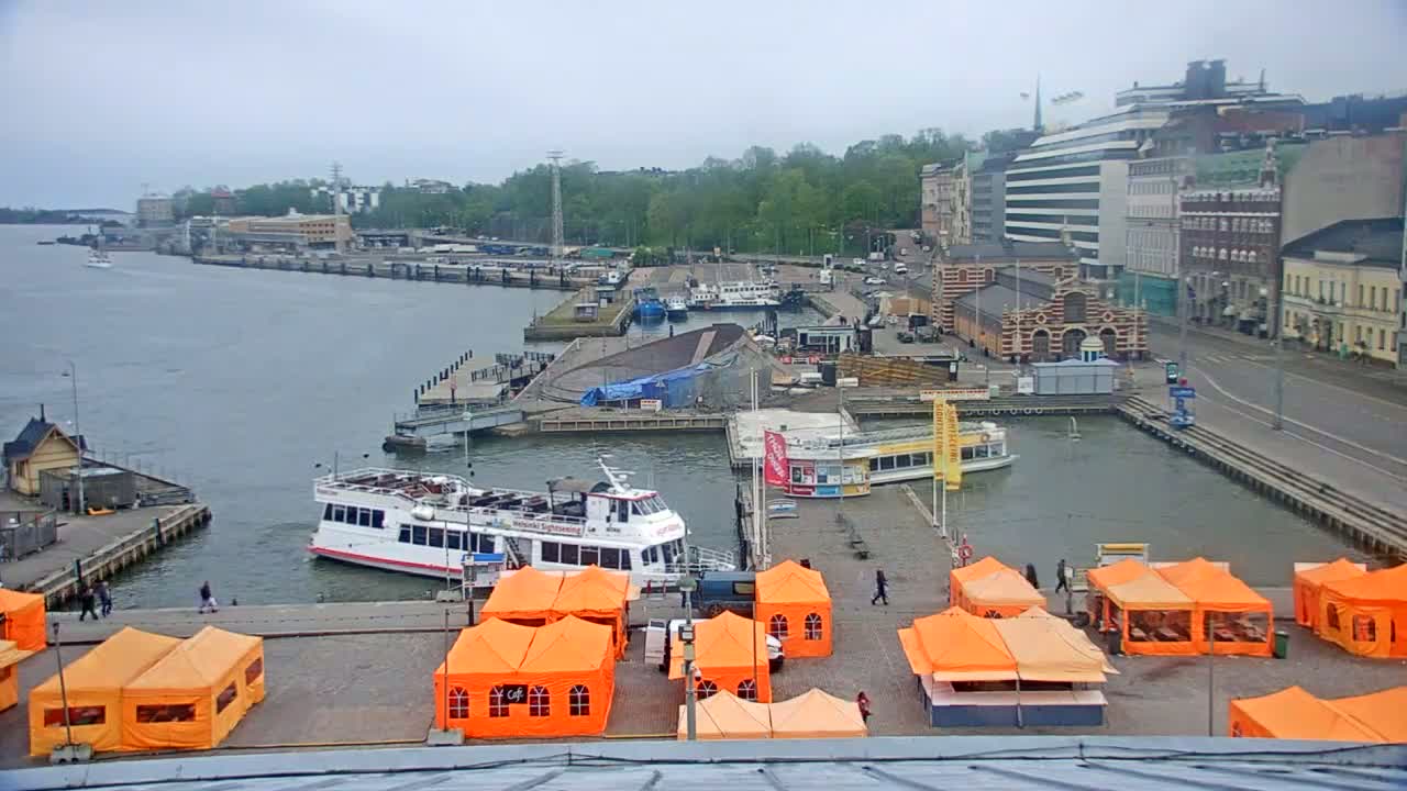 A harbor scene on an overcast day shows numerous boats docked near buildings and orange tents set up on a paved area.