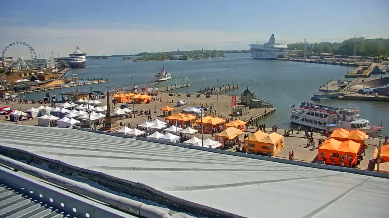 A harbor scene on a sunny day shows numerous market stalls, ferries, and a Ferris wheel, all near a body of water.