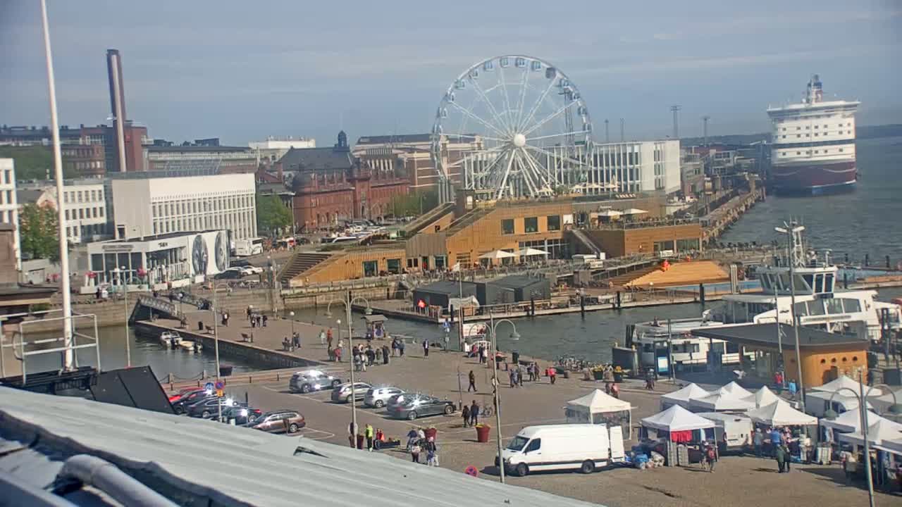 A harbor scene on a sunny day shows a Ferris wheel, buildings, docked boats, and people walking around a waterfront area.