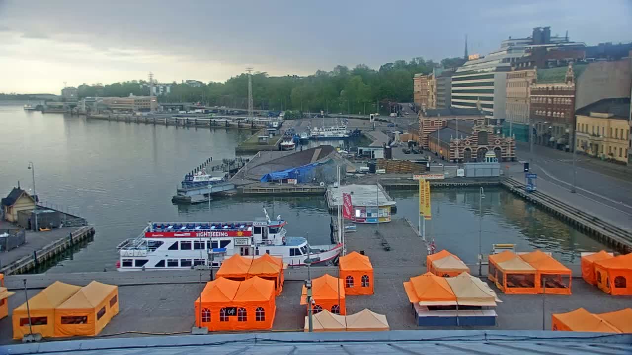 A harbor scene on an overcast day shows numerous boats docked near buildings and several orange tents set up on a paved area.