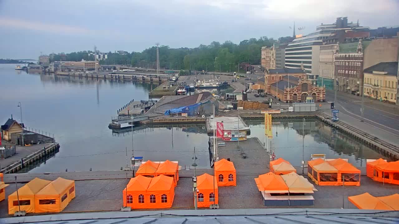 A harbor scene on an overcast day shows several buildings, boats, and orange tents situated along a waterfront with calm water.