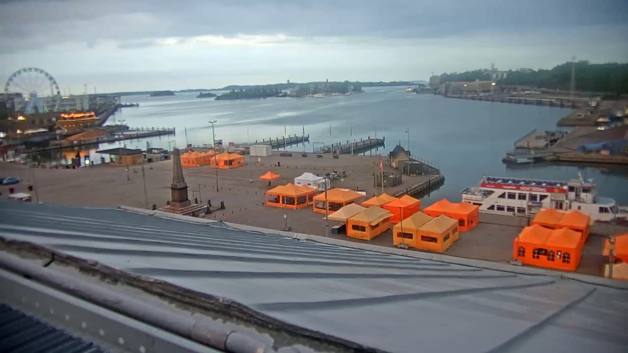 An overcast day reveals a harbor scene with numerous orange tents set up on a plaza next to a body of water, a Ferris wheel in the distance, and a docked passenger ship.